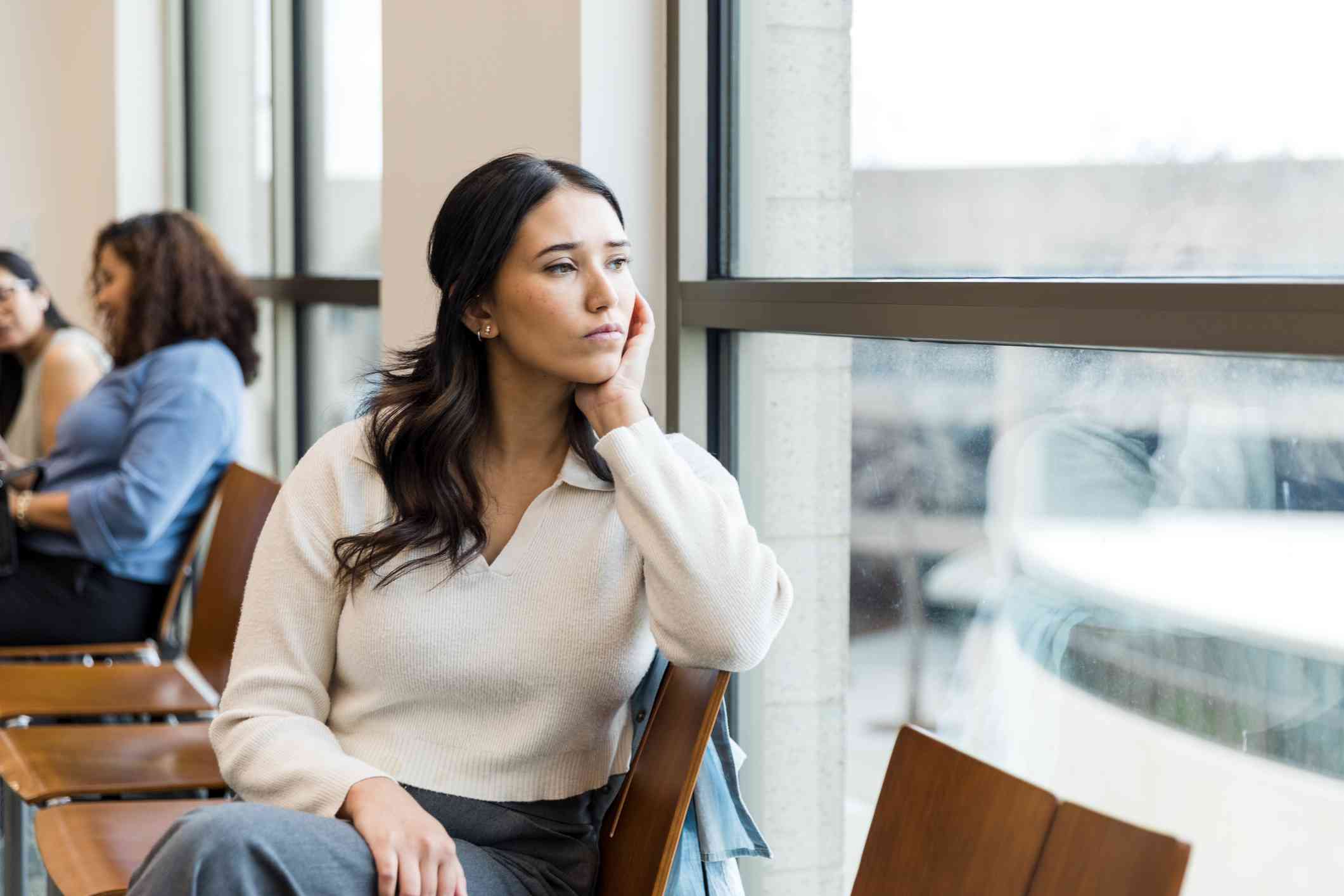 A woman in a tan shirt sits in achair away from the other people in the room and sadly gazes out of a window.