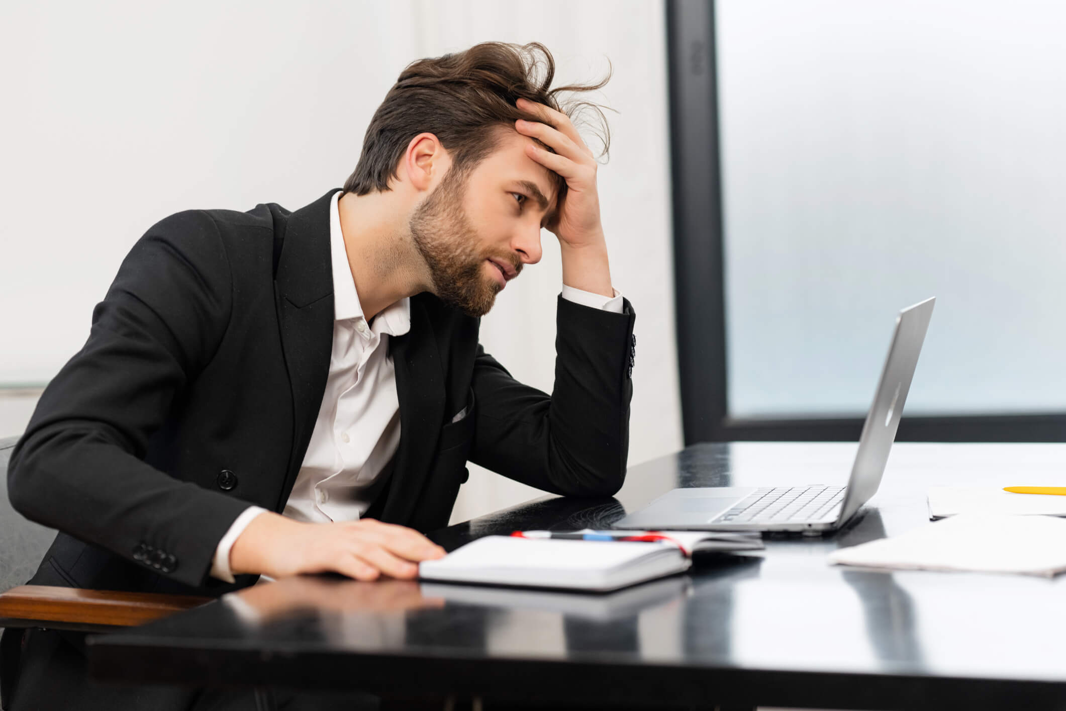 A man with brown hair and beard and a black blazer over a white collared shirt holds his head in frustration as he looks at his laptop screen and wonders how to fix his lack of motivation